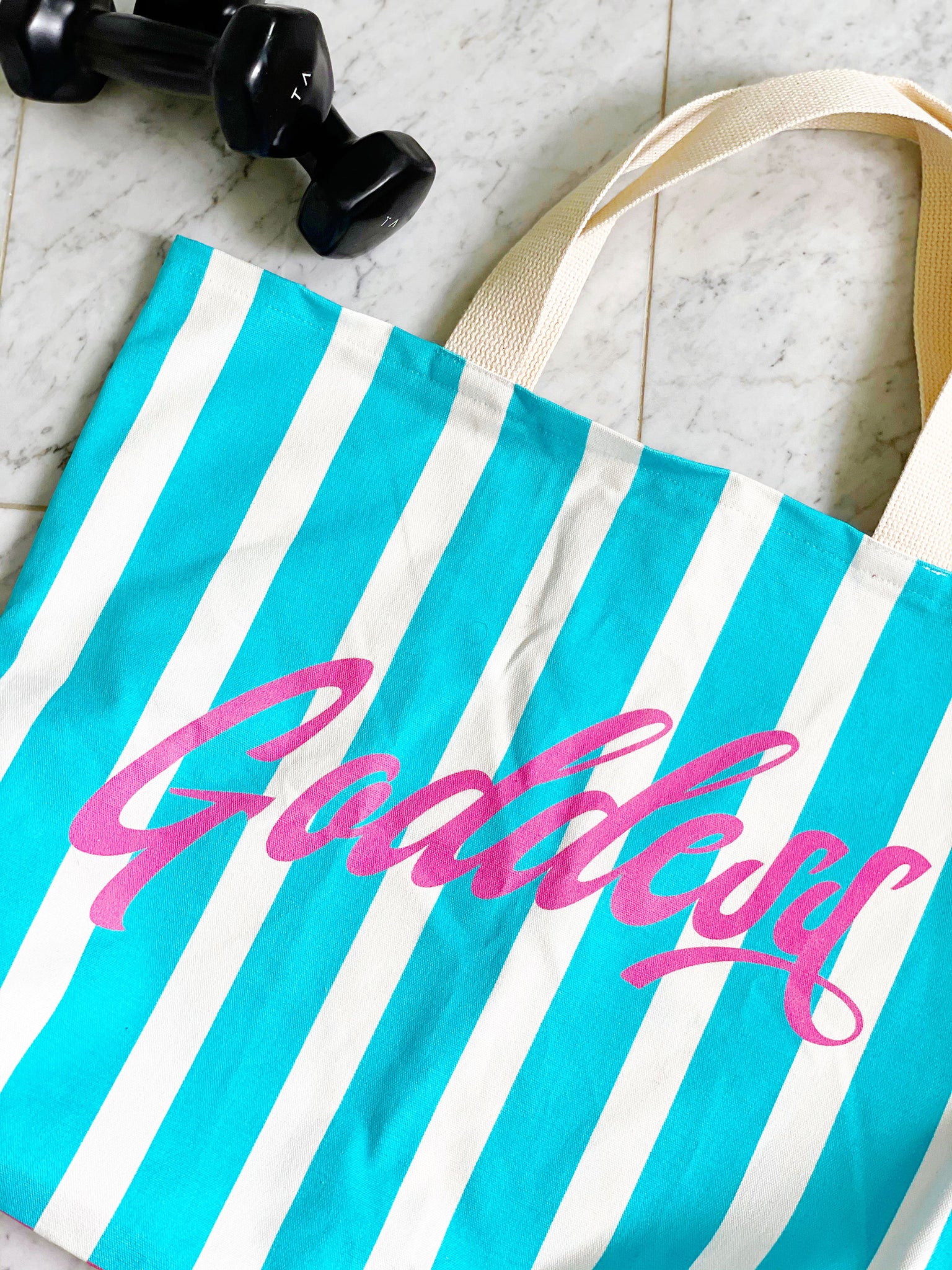 Our signature stripes luxe tote shown in a bold pink and blue with the word "Goddess" on the bag, features 2 interior pockets and a navy blue lining.